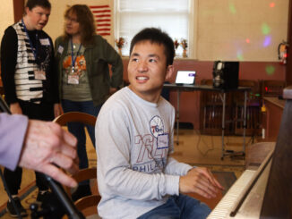 A man plays a piano with others listening in the background. He is smiling.