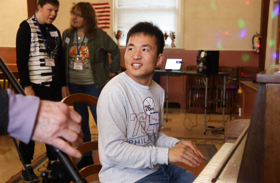 A man plays a piano with others listening in the background. He is smiling.