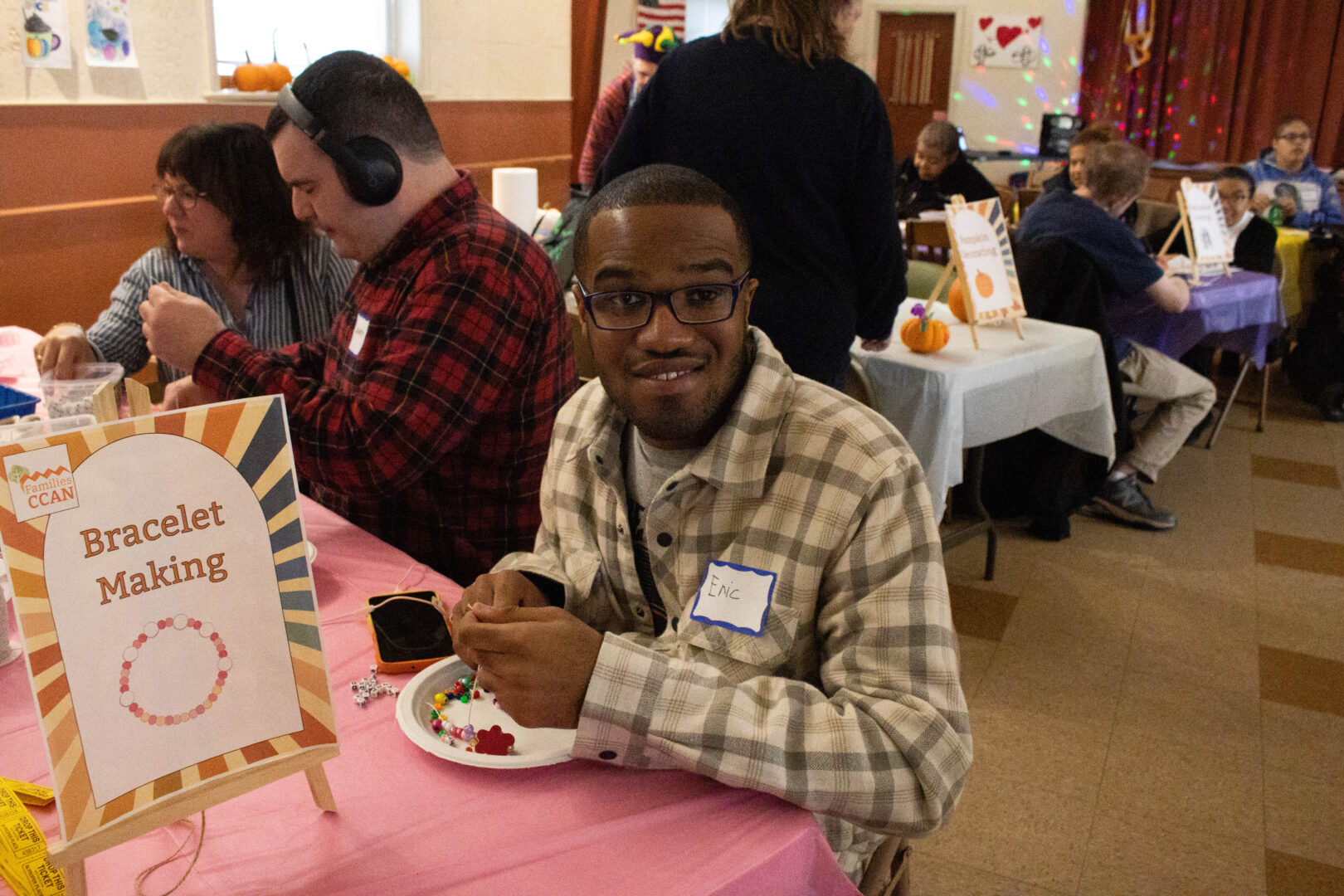 A man sits at a bracelet-making table, smiling as he strings colorful beads onto jewelry. A sign reading “Bracelet Making” stands on the table, and other people work on crafts at tables in the background during a busy fall festival.