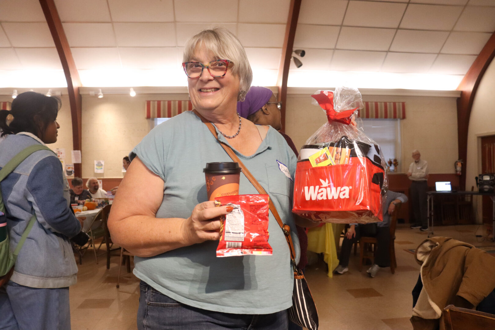 A smiling woman holds a Wawa-branded raffle basket along with a cup of coffee and a snack inside a busy community hall during a fall festival, with other attendees and tables visible in the background.