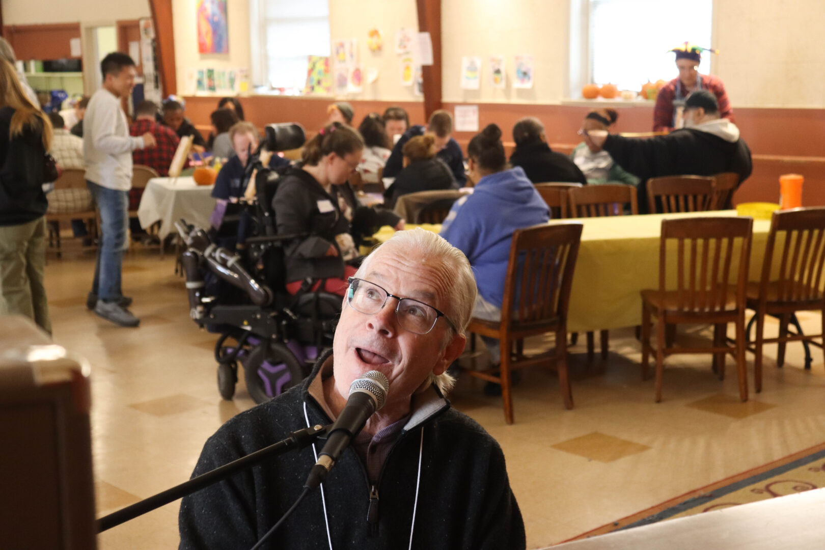 A man sings into a microphone while playing the piano at a fall festival, with attendees seated at long tables behind him participating in activities inside a community hall.