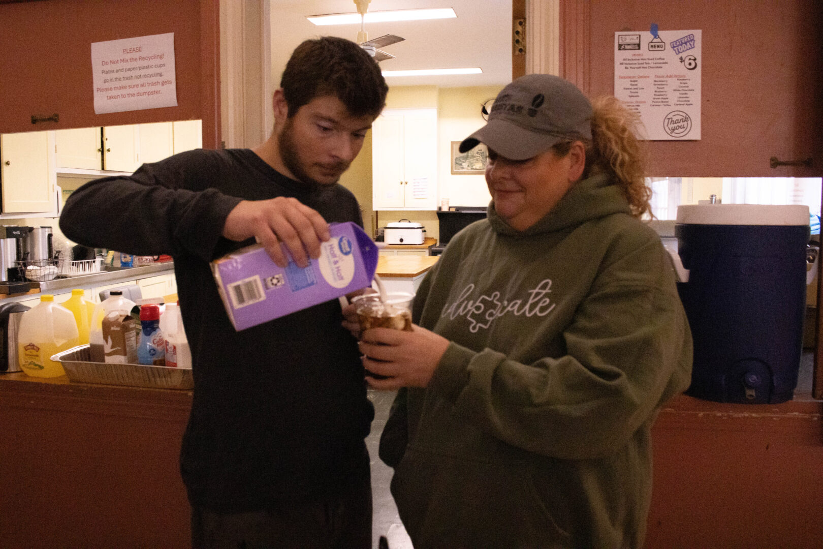 Two people stand at a refreshment counter as one pours cream into a cup of iced coffee held by the other, with coffee supplies and drink containers visible behind them inside a community kitchen.