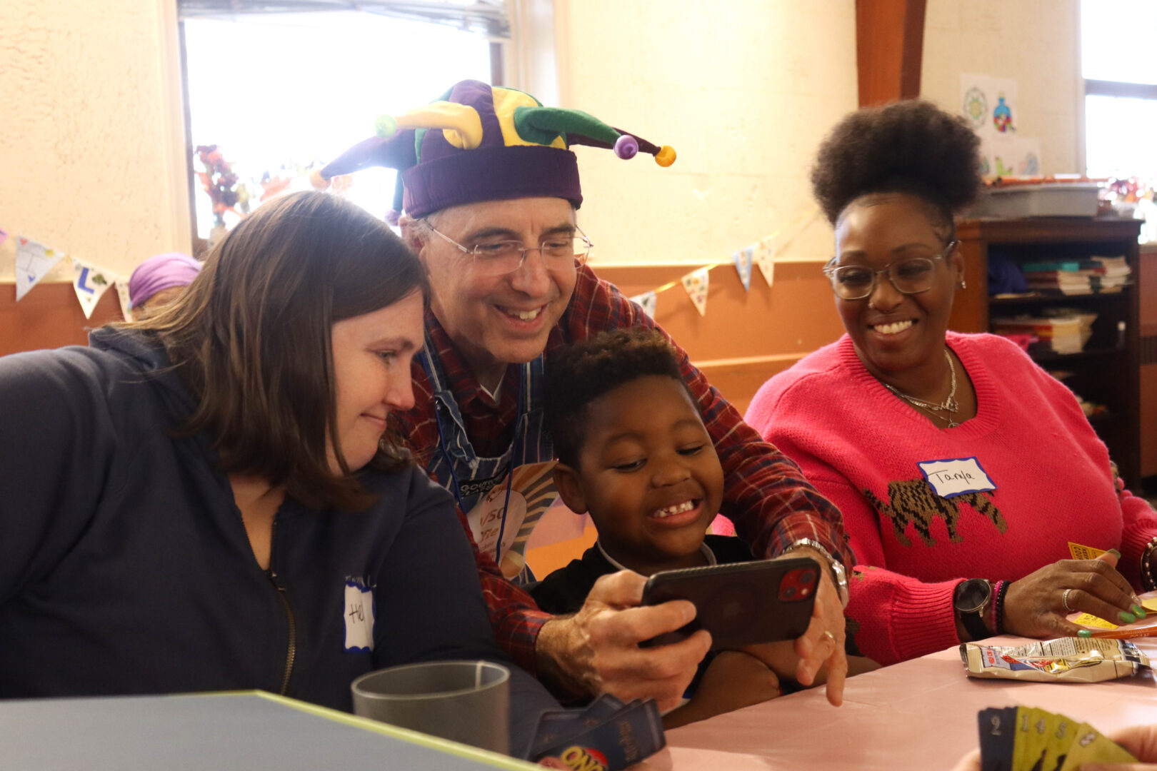 A man wearing a colorful jester hat shows something on his phone to a smiling child seated between two women at a table, as they gather together during a fall festival activity.