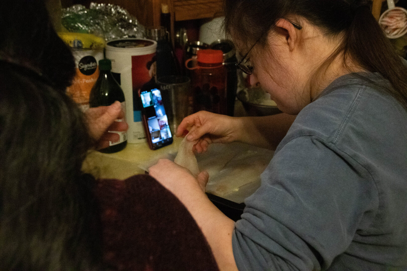 A woman prepares fish at a kitchen counter while following along on a smartphone set up for a video call, with cooking ingredients and utensils spread out around her.