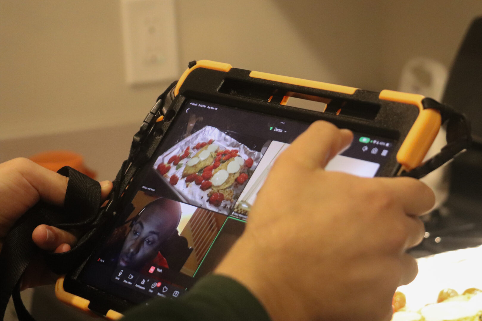 A person holds a tablet in a rugged orange case showing a Zoom meeting with a sheet pan of chicken parmesan.