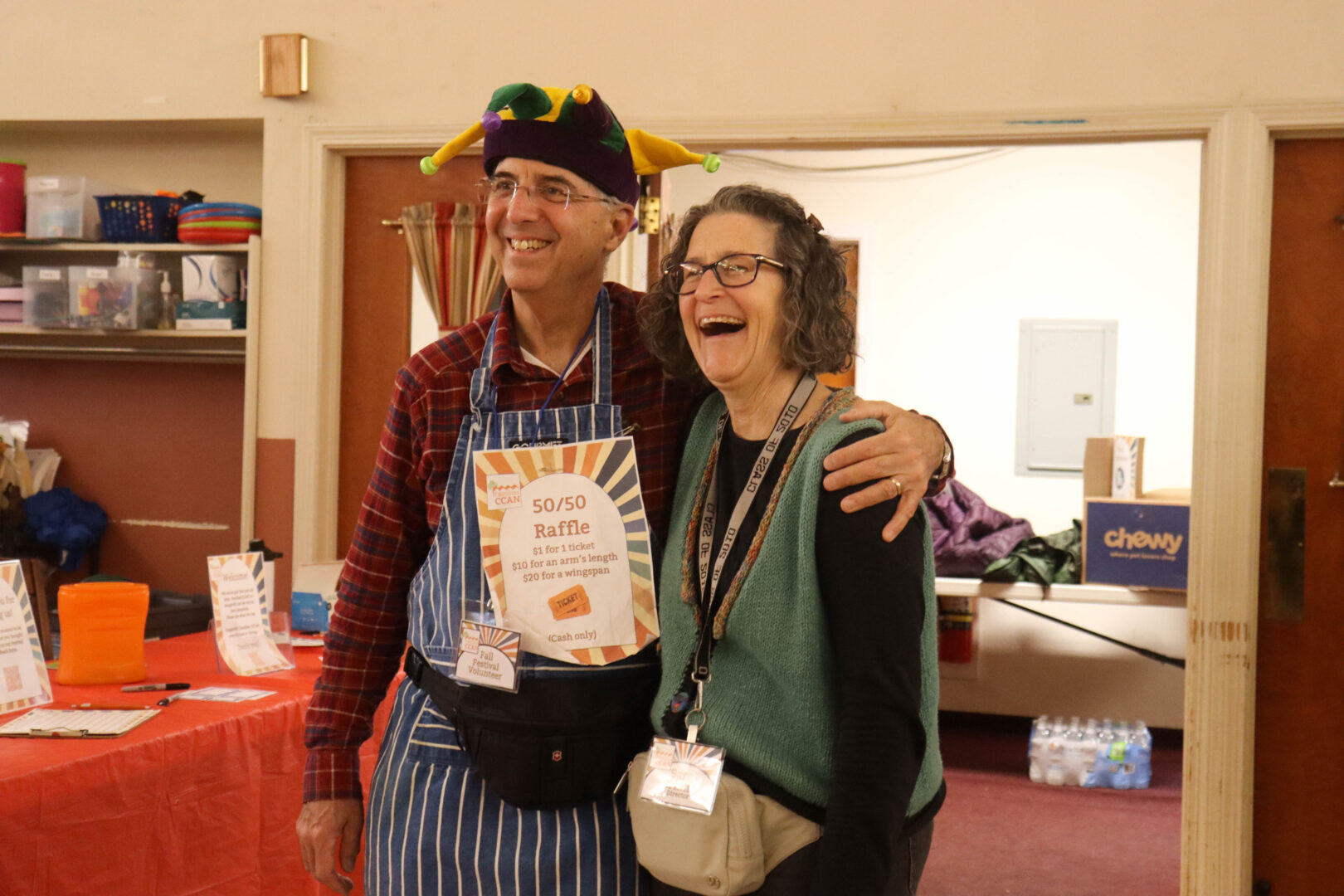 Sara and Allan stand together in a room with a red-clothed table and a "50/50 Raffle" sign. Allan wears a colorful purple, yellow, and green jester hat and a striped blue apron, while Sara wears glasses and a teal knit vest. They are both laughing and looking off-camera.