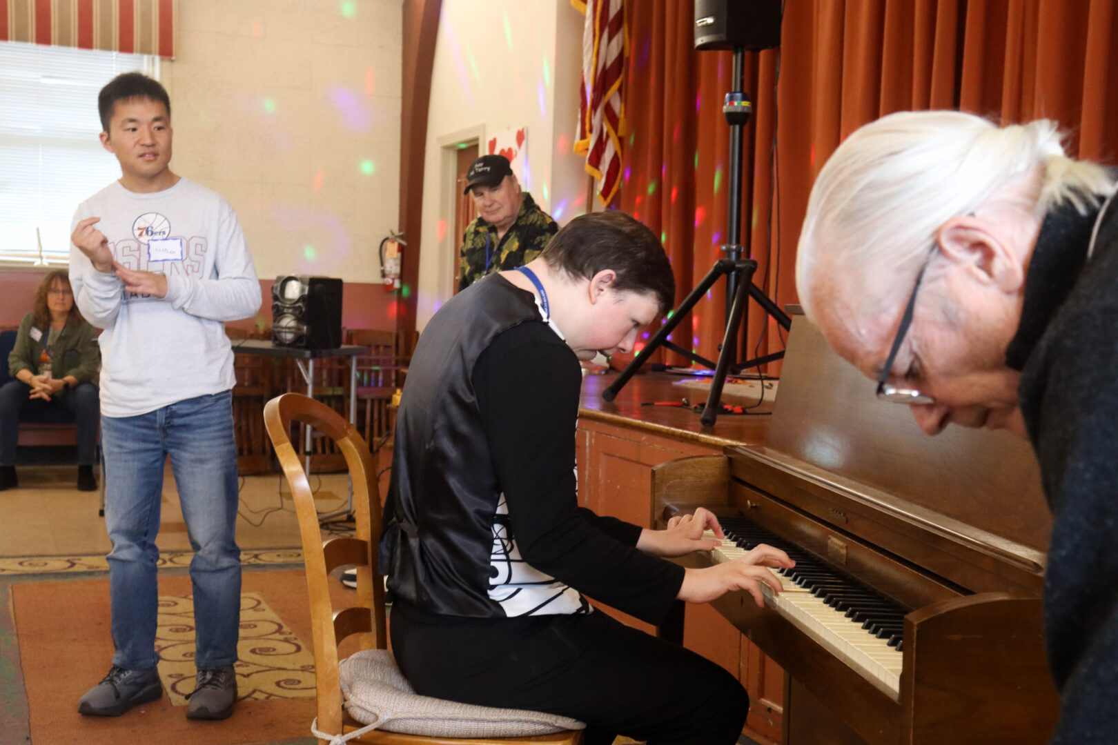 A young man playing an upright piano while another young man stands and dances in the background.