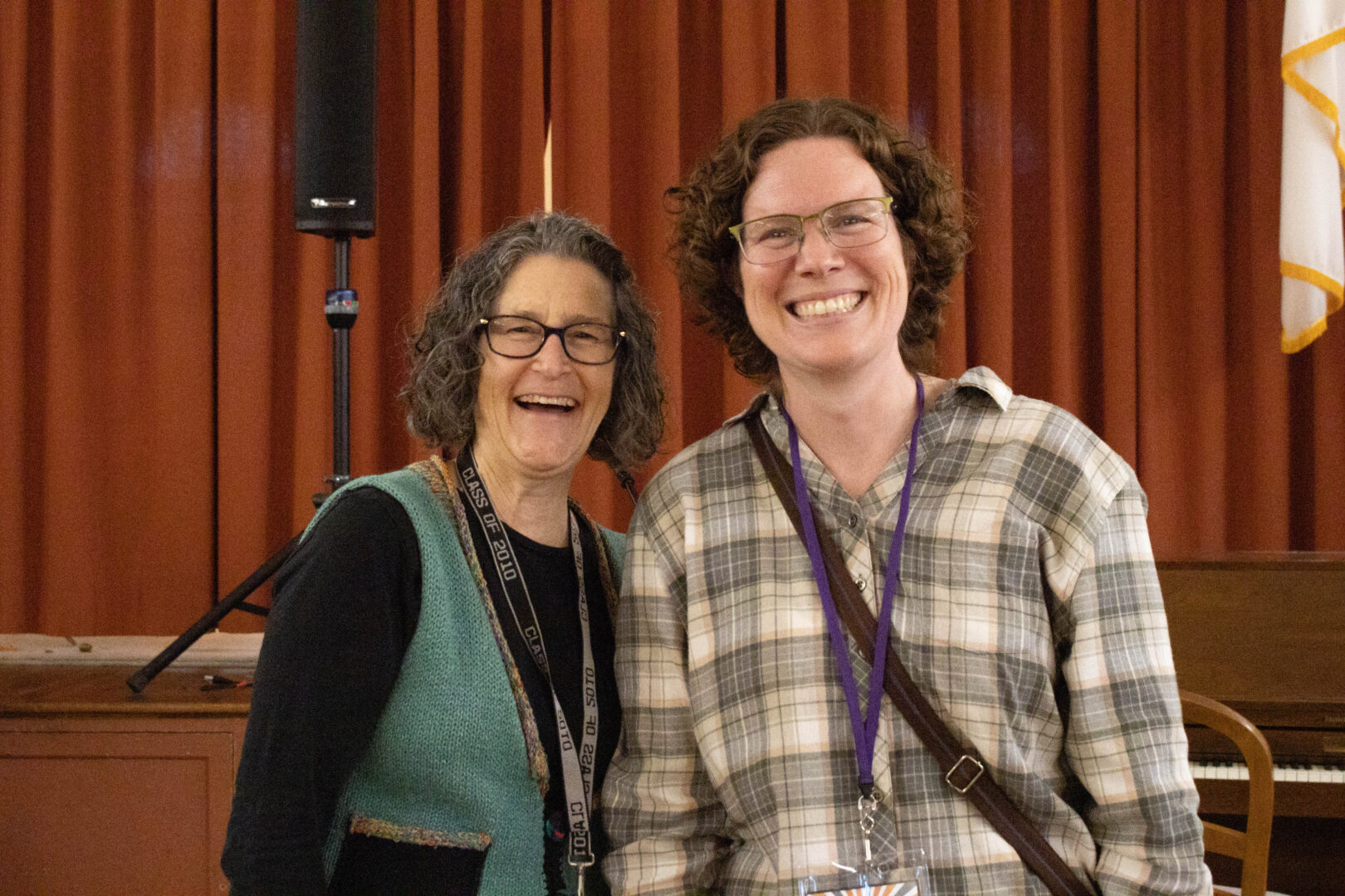 Sara, wearing glasses and a teal knit vest, and Abby, wearing glasses and a tan plaid shirt, stand side-by-side smiling for a photo. They are in a room with a wooden stage and orange curtains in the background.