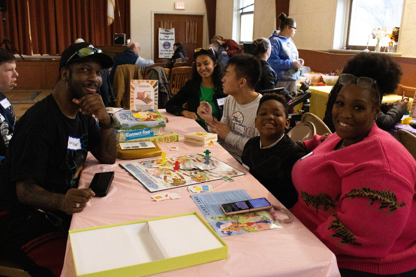 Tanya, wearing a pink sweater, and Arthur, in a black t-shirt and baseball cap, smile for a photo while seated at a pink-clothed table. They are joined by several others playing Candy Land, with a stack of other games like Connect Four nearby. The room is filled with other attendees and yellow-covered tables in the background.