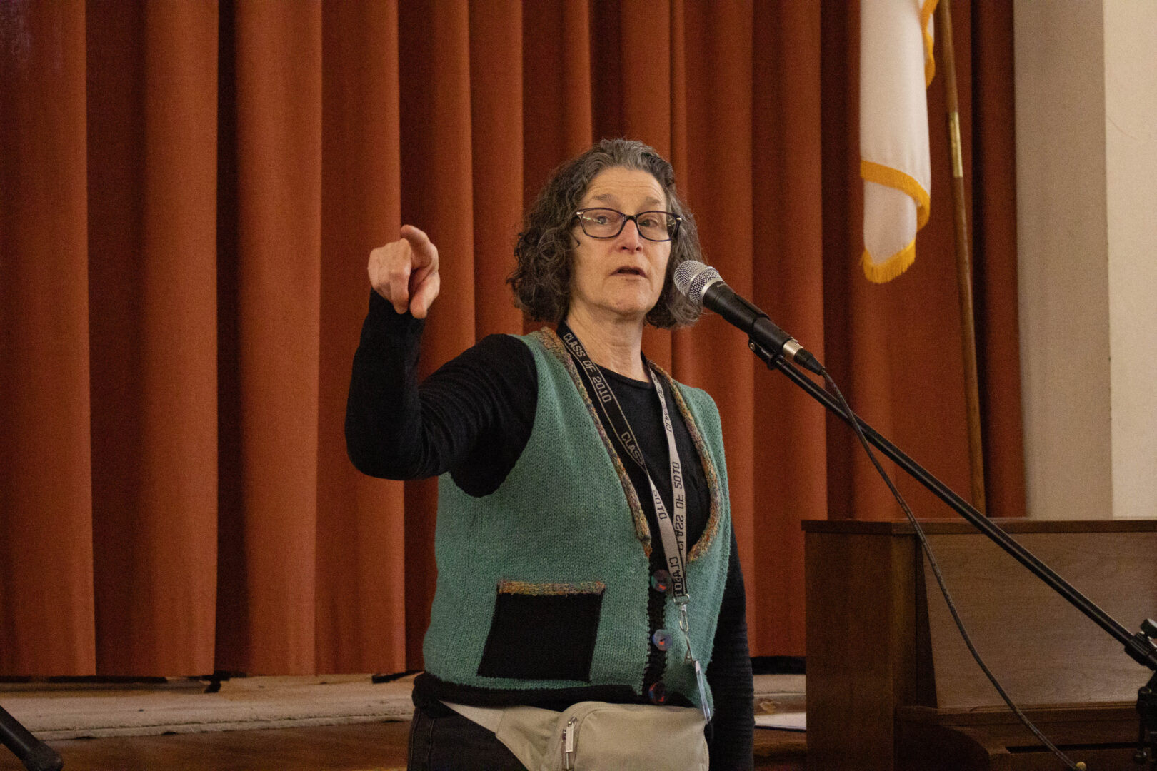 A woman, Sara, speaking into a microphone on a stage while gesturing with her hand.