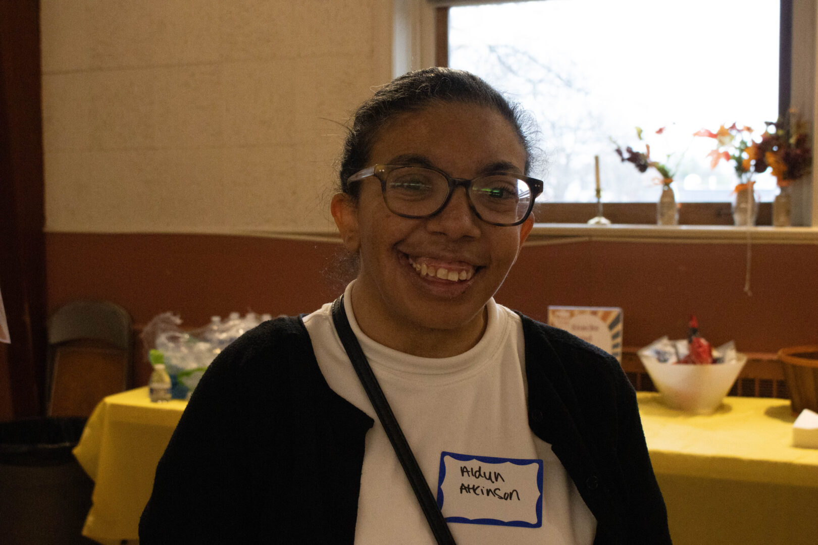 Aldyn, a young woman with dark hair pulled back and wearing glasses, smiles warmly for a portrait. She is wearing a white t-shirt with a blue-bordered name tag that says "Aldyn Atkinson" and a black cardigan. In the background, a table with a yellow tablecloth holds snacks and water bottles, and a window with fall floral decorations is visible.