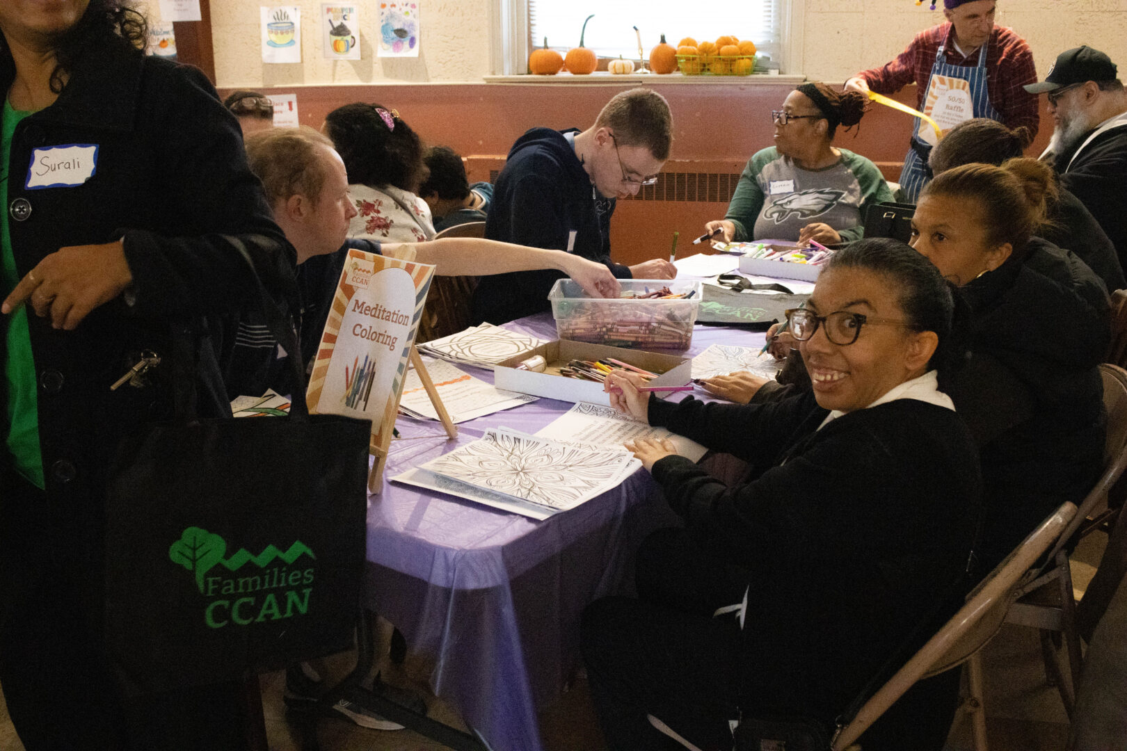 Aldyn, a young woman with dark hair and glasses, sits at a table and smiles toward the camera while holding a colored pencil. A sign on the table reads "Meditation Coloring" next to containers of crayons and various coloring sheets. In the background, other attendees are also coloring, and a man in a jester hat is visible near a window.