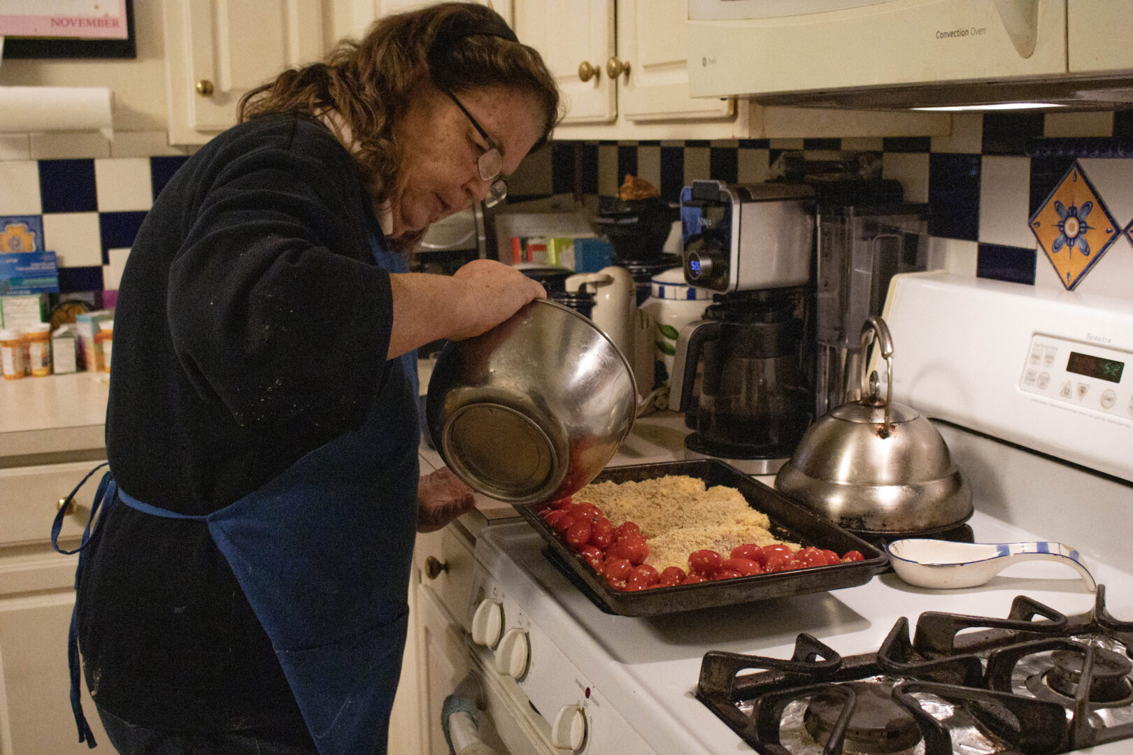 In a kitchen with a blue-and-white tiled backsplash, Mary, wearing a blue apron and glasses, tilts a stainless steel bowl to pour red cherry tomatoes onto a black baking tray. The tray already contains breaded chicken cutlets prepared for a sheet pan meal. A silver kettle and a coffee maker are visible on the counter in the background.