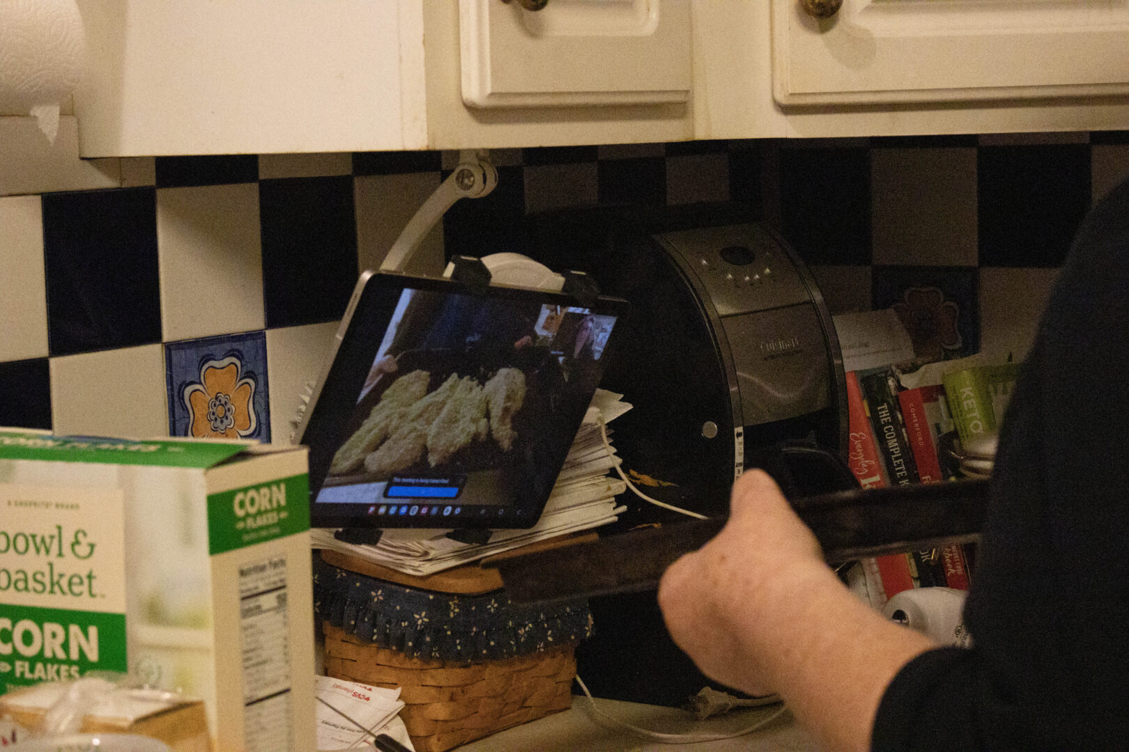 A hand holds up a baking tray toward a tablet displaying a virtual Zoom cooking class in a kitchen.