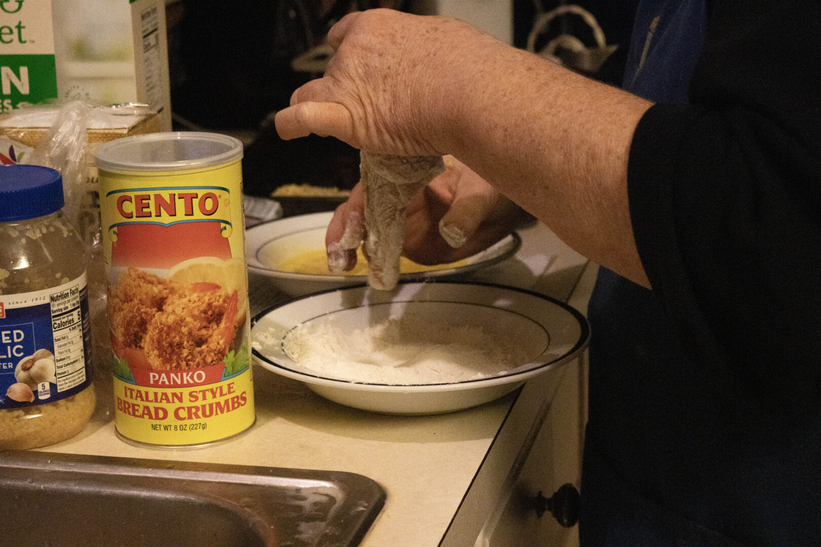 A person prepares a breading station on a kitchen counter. Their hands are shown dipping a piece of raw chicken into a white bowl of flour, with a second bowl of egg wash visible behind it. Next to the bowls is a yellow container of Cento Panko Italian Style Bread Crumbs and a jar of minced garlic.