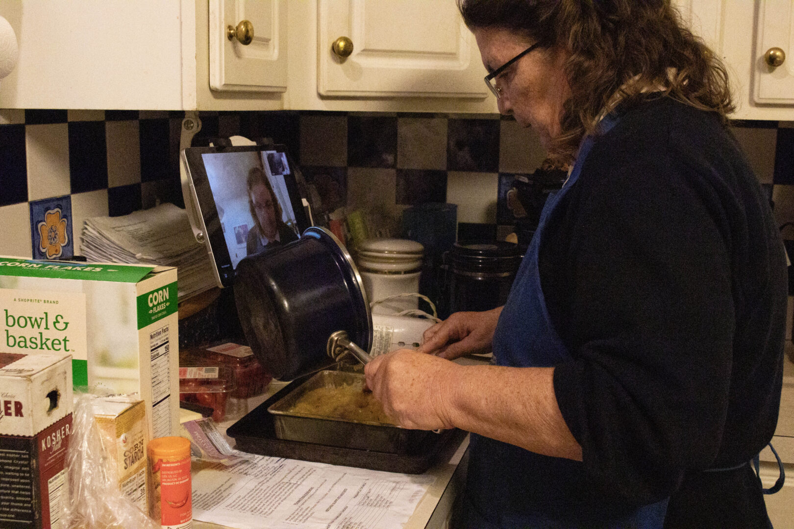 A side-profile view of Mary, wearing a blue apron, tilting a small dark blue saucepan to pour a yellow pineapple mixture into a silver baking pan. On her kitchen counter, a tablet propped against the wall displays a Zoom meeting with another participant's face visible. Boxes of corn flakes, corn starch, and other baking ingredients surround the workspace.