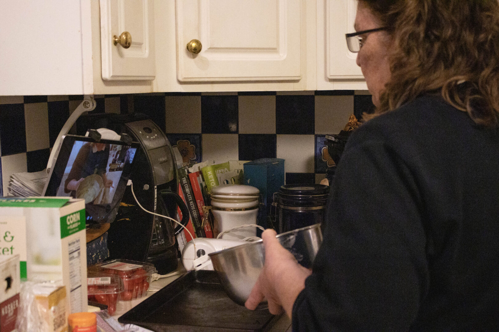 Mary holds a metal mixing bowl over a baking tray while her kitchen workspace and a tablet showing a virtual class are visible.