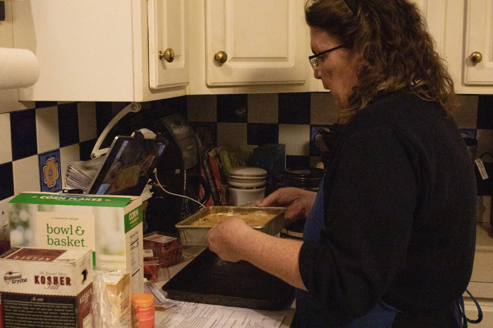 A side-profile view of Mary, wearing a blue apron, holding a silver square baking pan filled with a yellow-colored pineapple mixture. Her kitchen counter is crowded with ingredients like "Corn Flakes," "Kosher Salt," and "Corn Starch," while a tablet propped in the background shows a Zoom meeting.