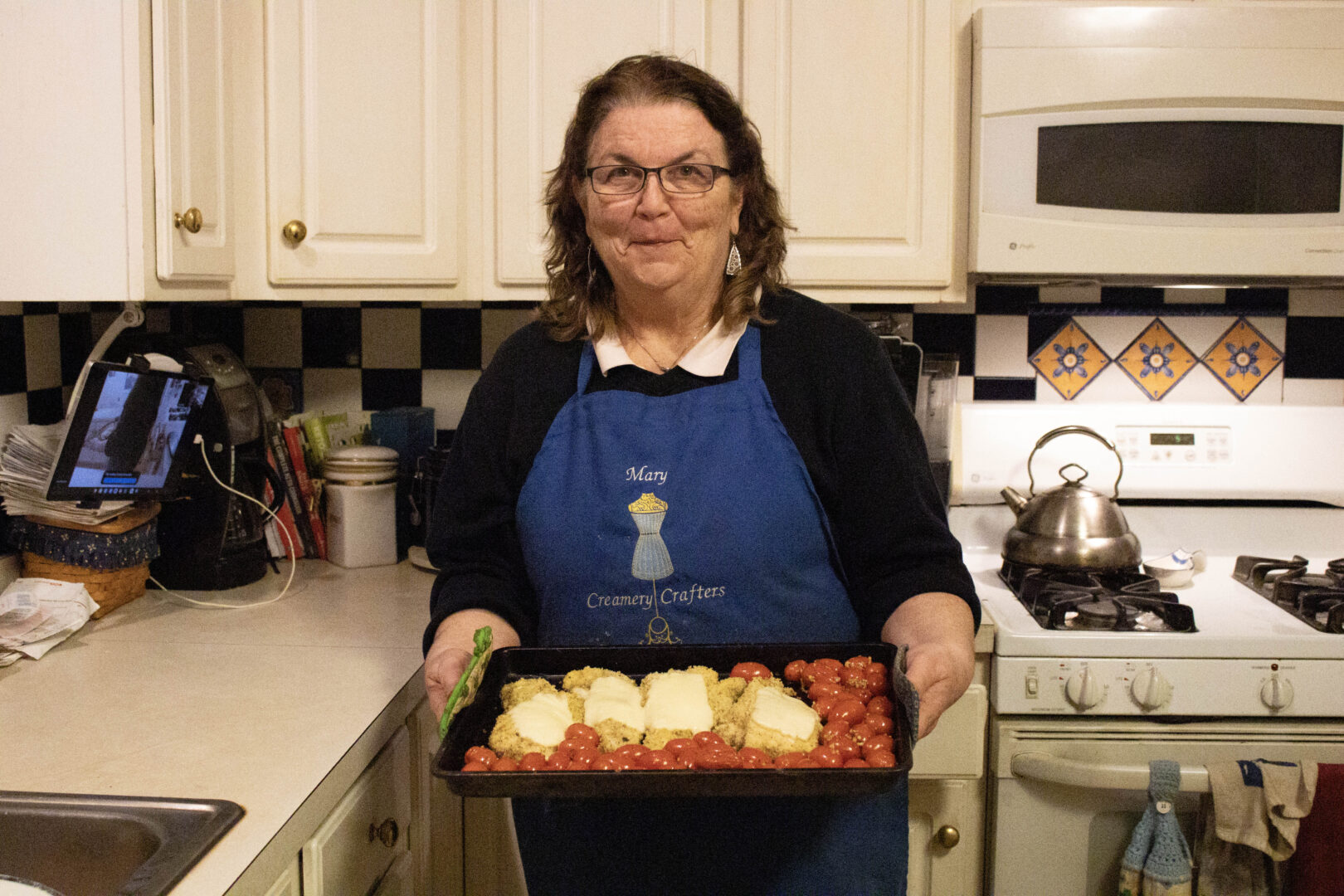 Mary holds up a completed sheet pan of chicken parmesan and roasted tomatoes in her kitchen.