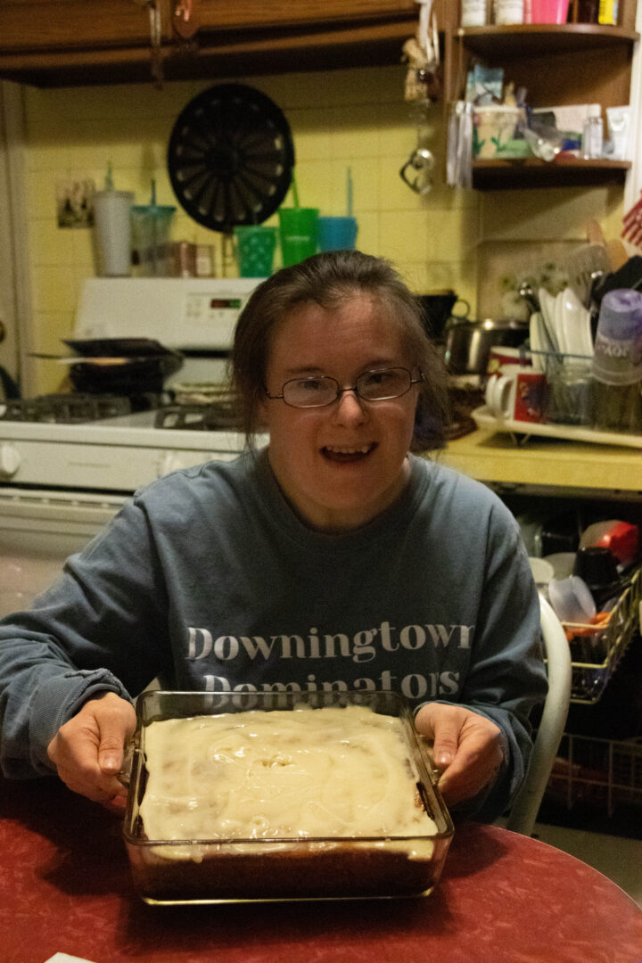 A smiling young woman holds a glass baking dish containing a frosted dessert in her kitchen.