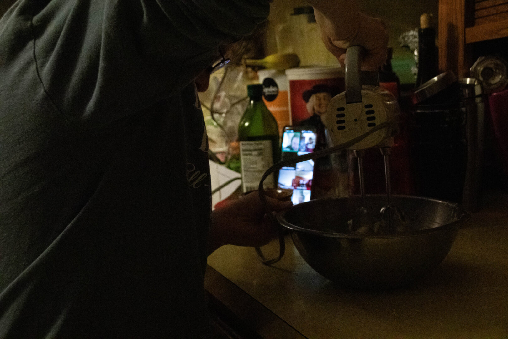 A close-up, side-profile view shows Aubrey’s arm and torso as she holds a white hand mixer over a stainless steel mixing bowl. On the kitchen counter behind the bowl, a smartphone is propped up, displaying a Zoom meeting with multiple participant tiles. Various kitchen items, including a bottle of olive oil and a container of oats, are visible in the dimly lit background.