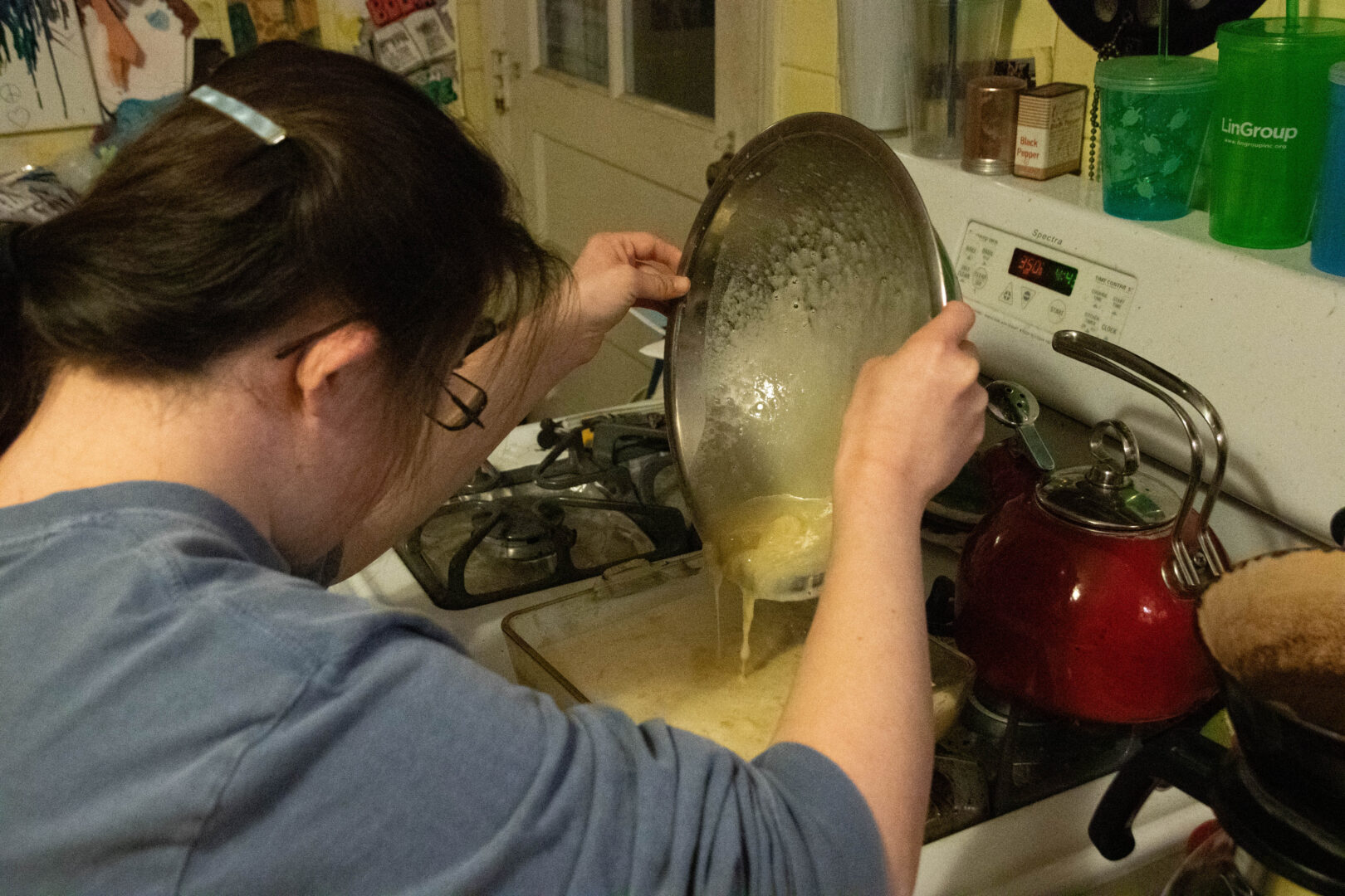 Aubrey, seen from behind with her dark hair pulled back, tilts a stainless steel bowl with both hands to pour batter into a rectangular glass pan on her stove. A red tea kettle and various kitchen items are visible on the counter and shelves in the background.