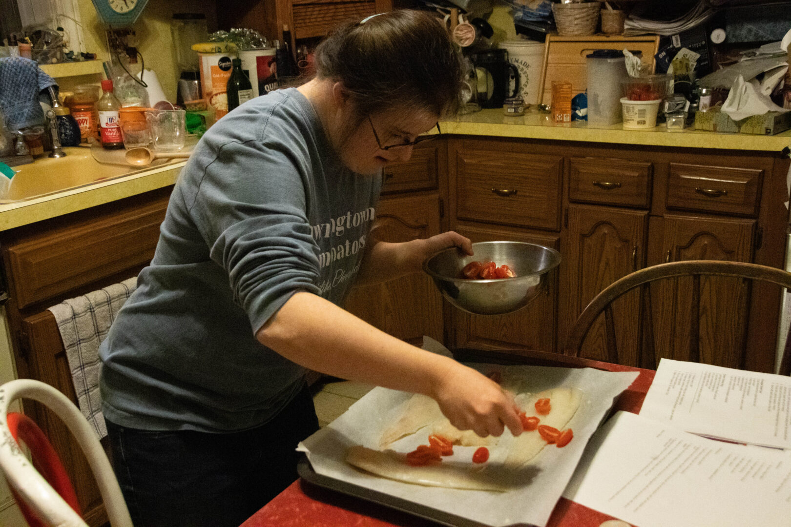 Aubrey, wearing glasses and a blue "Downingtown Dominators" sweatshirt, stands at a red table and reaches into a metal bowl to sprinkle halved cherry tomatoes over white tilapia fillets. The baking sheet is lined with white parchment paper, and printed recipe sheets are visible on the table.