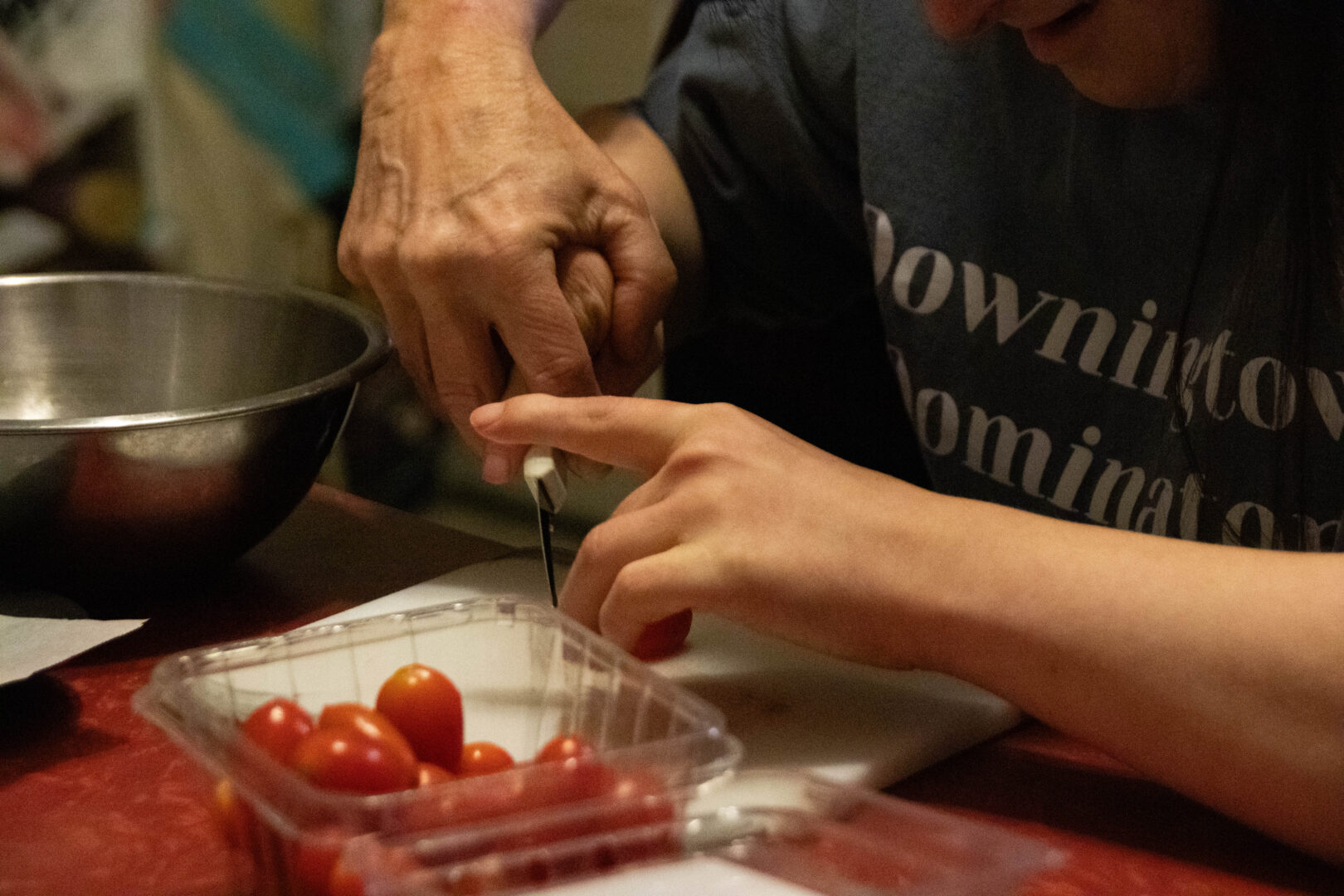 A close-up of two people's hands working together to slice cherry tomatoes on a small white cutting board.