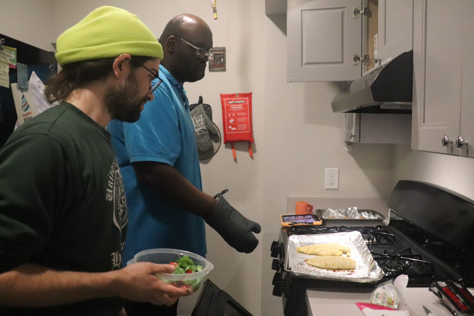 William, a man in a blue polo shirt and glasses, wearing an oven mitt, stands by a stove where a baking sheet with two breaded chicken cutlets rests on the burners. Next to him, another man in a green sweatshirt and a lime-green beanie holds a clear bowl of salad. A tablet propped up on the counter displays a Zoom meeting.