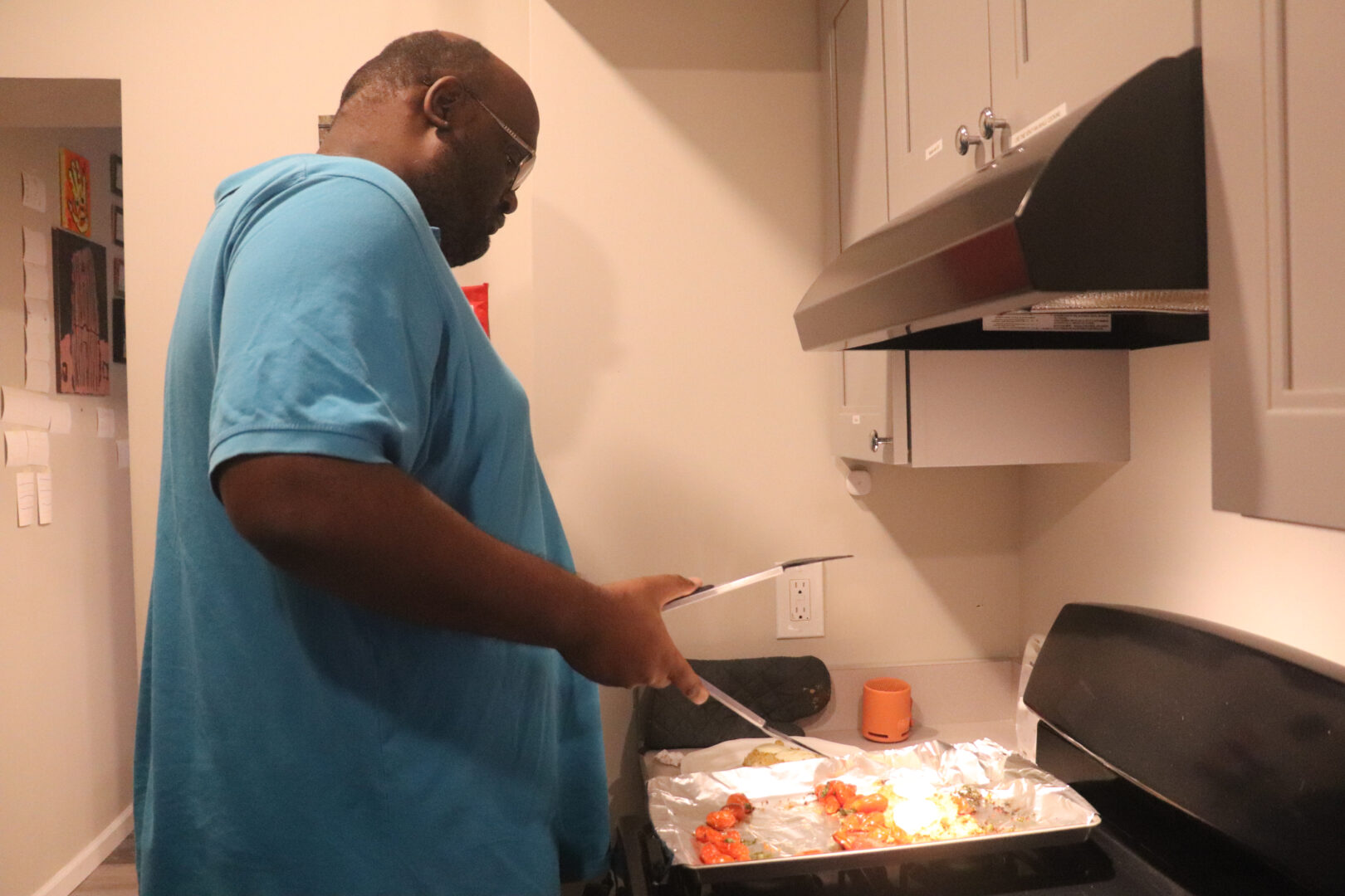 William, a man with glasses wearing a bright blue polo shirt, is shown in profile standing at a stove. He uses metal tongs to lift a piece of breaded chicken from a baking sheet lined with aluminum foil, which also contains several roasted cherry tomatoes. 