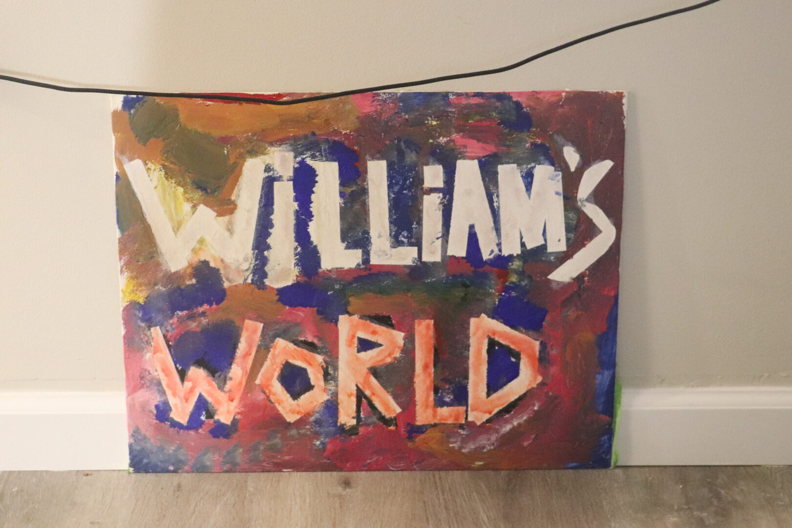 A rectangular canvas features the words "WILLIAM’S WORLD" painted in large, bold, block letters. The top word, "WILLIAM'S," is painted in white over a textured background of blue, red, and yellow. The word "WORLD" is painted in orange at the bottom. The canvas is propped up against a plain light-colored wall, resting on a light-brown wood-grain floor.