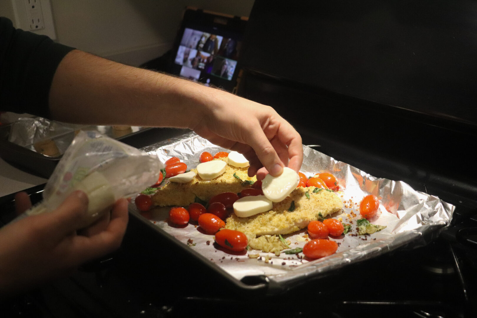 A close-up shows hands placing fresh mozzarella slices onto breaded chicken on a baking sheet while a tablet in the background displays a Zoom class.