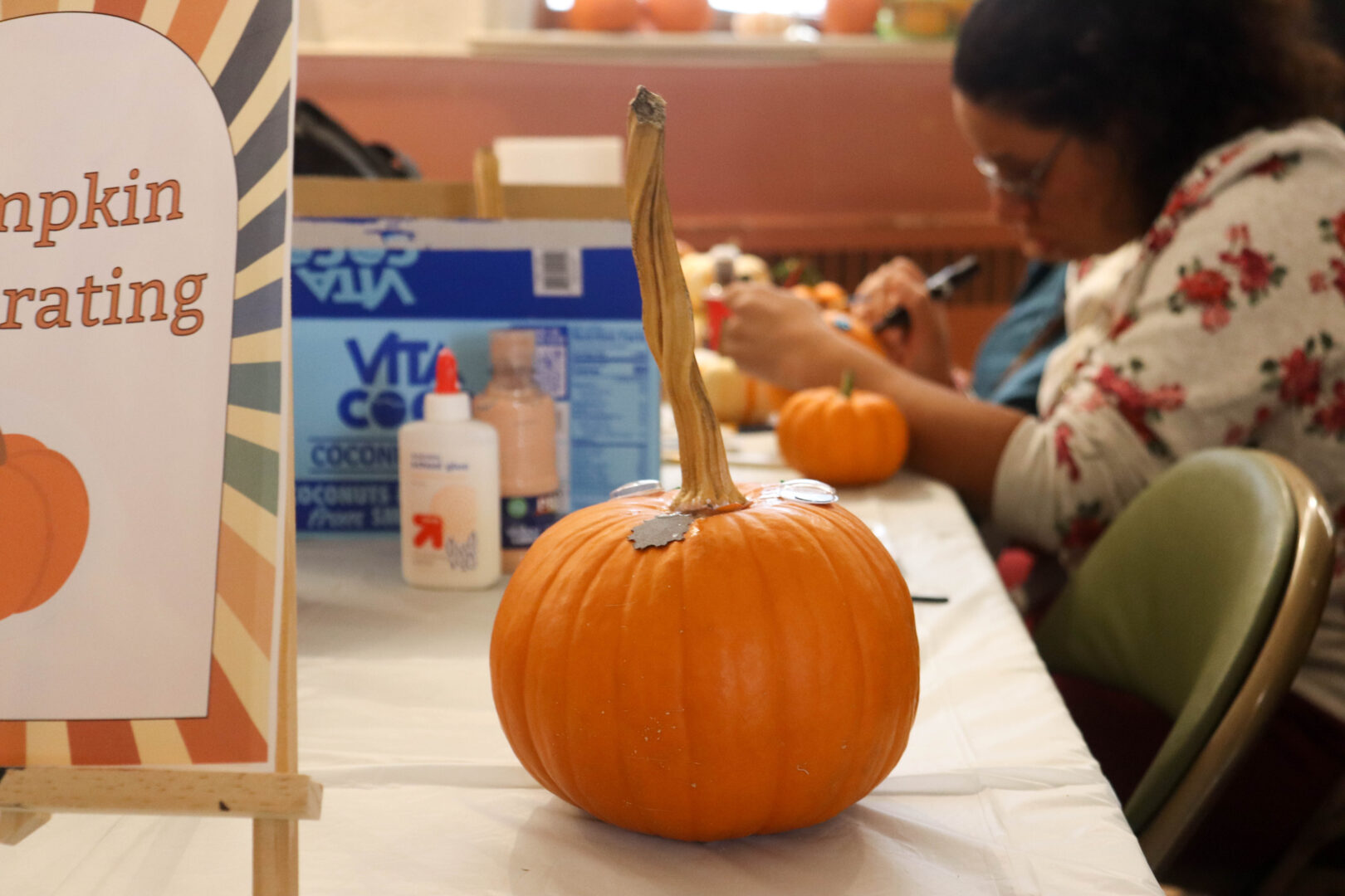 A small orange pumpkin sits on a table set up for decorating, with glue bottles and supplies nearby. In the background, a person concentrates on decorating another pumpkin at a Fall Fest event.