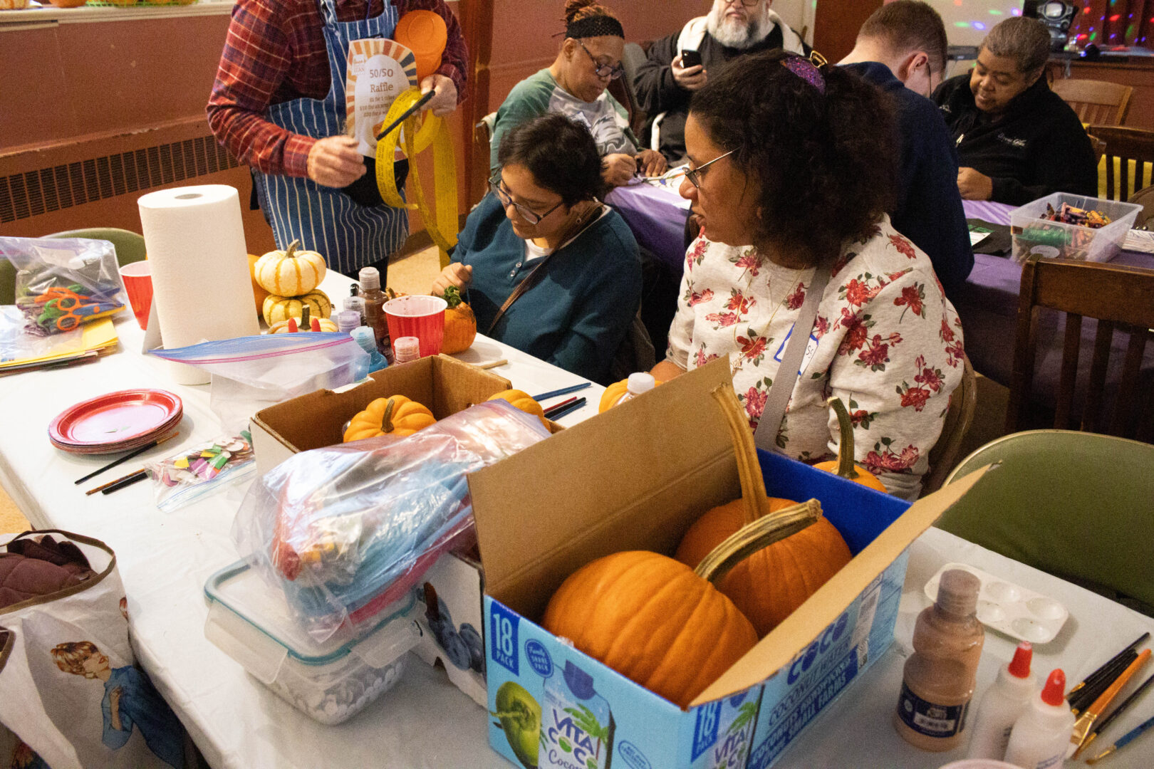 Two people sit at a pumpkin-decorating table, painting and coloring small pumpkins surrounded by craft supplies, while other festival participants work at tables in the background.