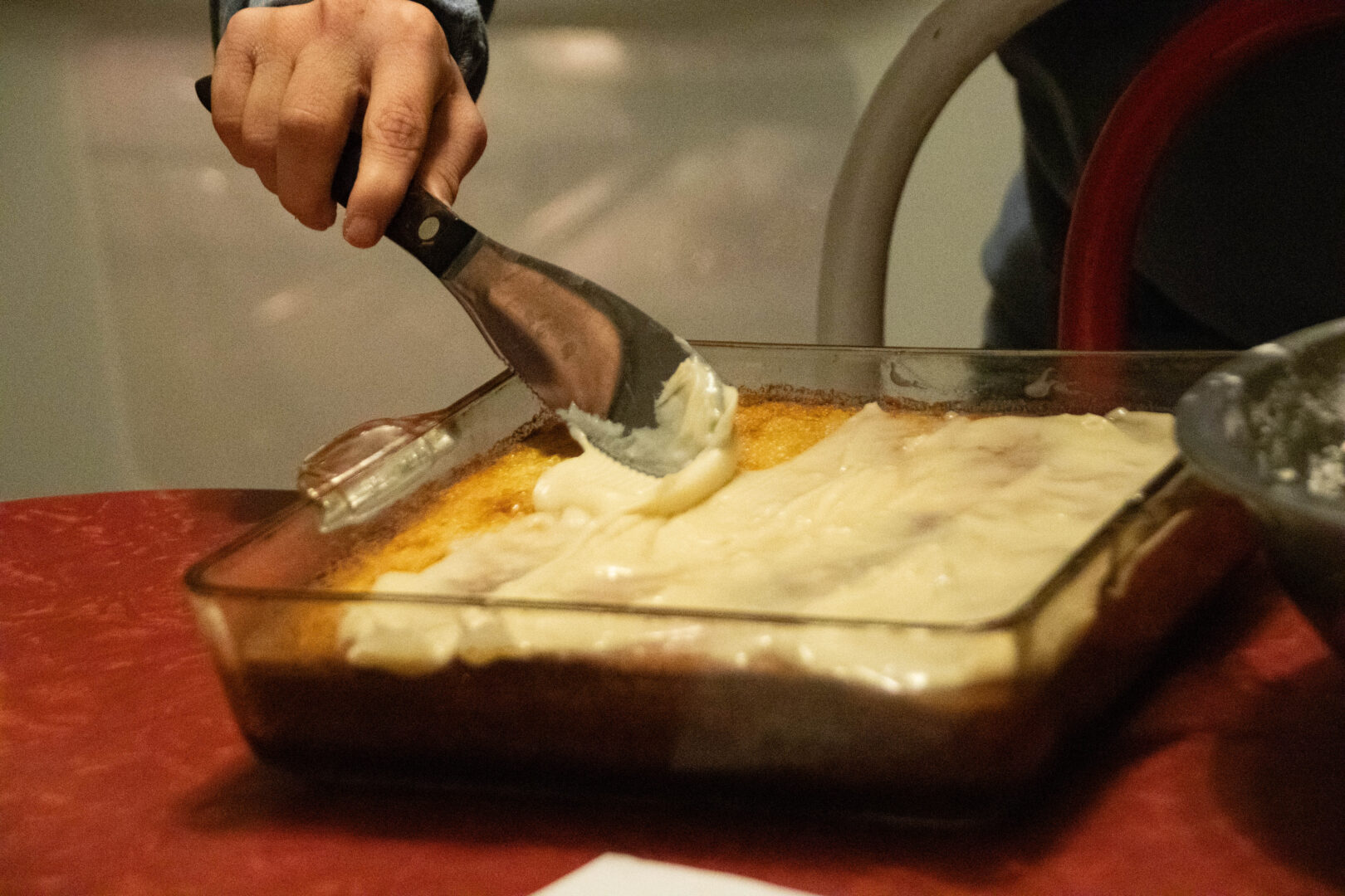 A close-up of a hand using a metal spatula to spread white frosting across a golden-brown dessert in a glass baking dish.