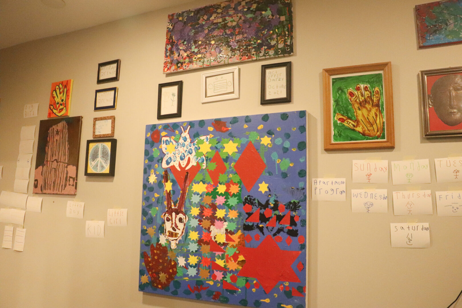 A tan wall is covered in an eclectic mix of artwork. A large central blue canvas features abstract shapes, yellow stars, and red diamonds. Surrounding it are smaller framed pieces, including a painting of a hand, a peace sign, and abstract sketches. To the right, several small white papers are taped to the wall, each featuring a day of the week and a small hand-drawn figure.
