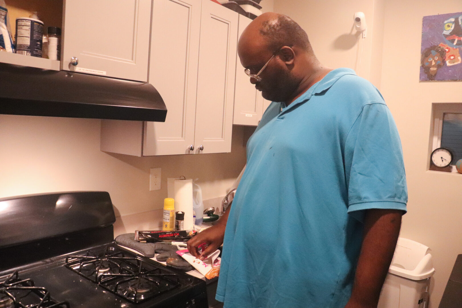 A side-profile view of William, a man with glasses wearing a bright blue polo shirt, standing in his kitchen. He is looking down at a counter next to a black gas stove, where cooking supplies like aluminum foil, paper towels, and various ingredient packages are visible. The kitchen has light grey cabinets and a white wall.